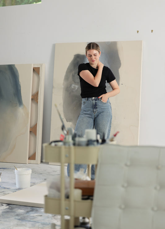 Woman standing in a bright studio beside large abstract beige and blue paintings with a paint cart in the foreground