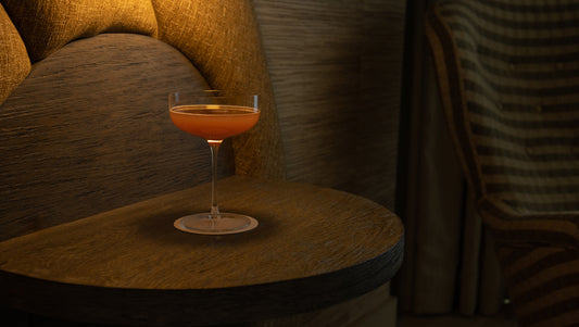 Single Fable coupe glass with an amber cocktail on a dark wood side table in a dimly lit lounge