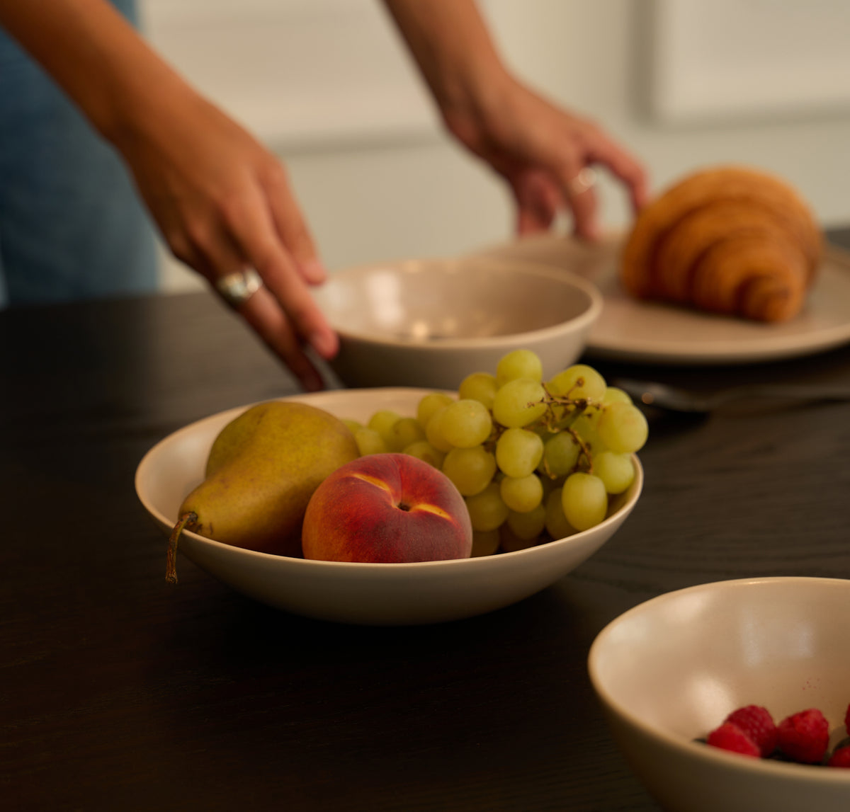 Hands setting beige Fable stoneware bowls of fresh fruit and a croissant on a dark wood dining table