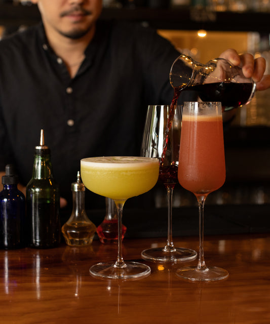 Bartender pouring a cocktail into a Fable champagne flute beside a wine glass and coupe on a wooden bar