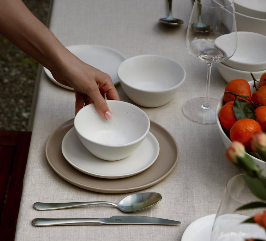 Hand placing a Fable speckled white bowl on a beige dinner plate at an outdoor table with oranges and wine glasses