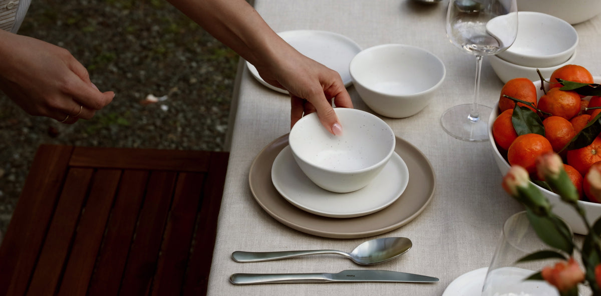 Hand placing a white Fable stoneware bowl on a plate at an outdoor table with citrus and wine glass