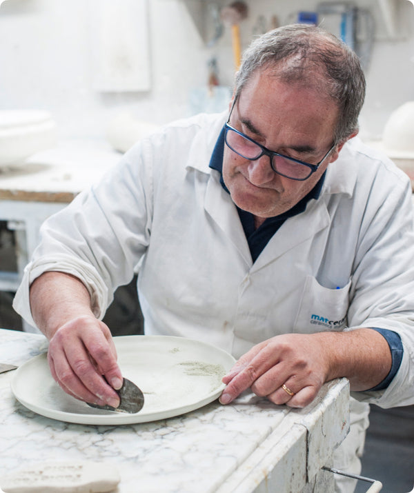 Ceramic artisan in a white coat finishing a Fable plate by hand at a workshop bench