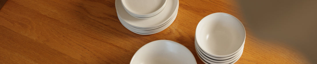 White Fable plates and bowls arranged in stacks on a warm wood surface in natural light