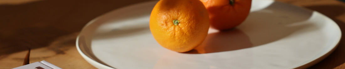 Two oranges resting on a white ceramic serving plate on a wooden table in warm light