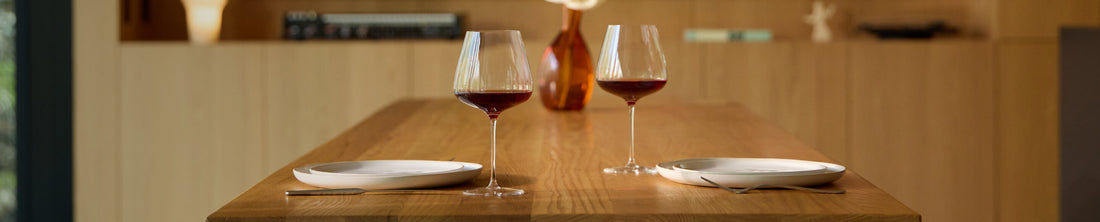 Two red wine glasses and white plates on a warm oak dining table in a modern kitchen