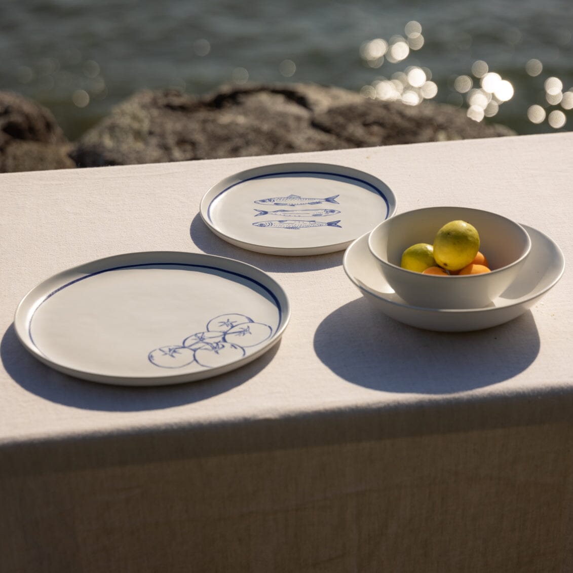 Ceramic plates with blue designs and a bowl of fruit on a stone surface near water.