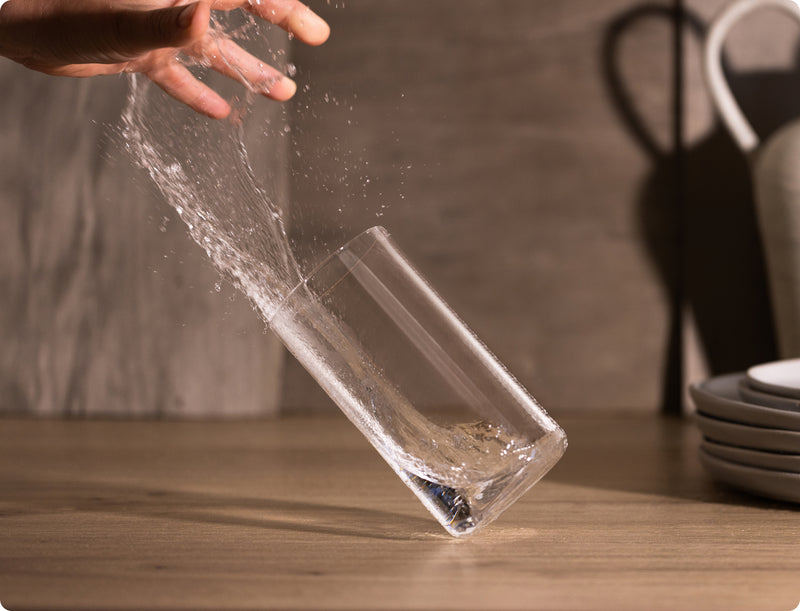 Hand splashing water from a Fable clear glass tumbler over a wooden table with stacked plates