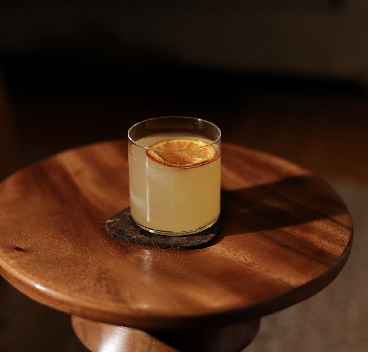 Fable clear glass tumbler holding a citrus cocktail with a dried orange slice on a round wooden side table