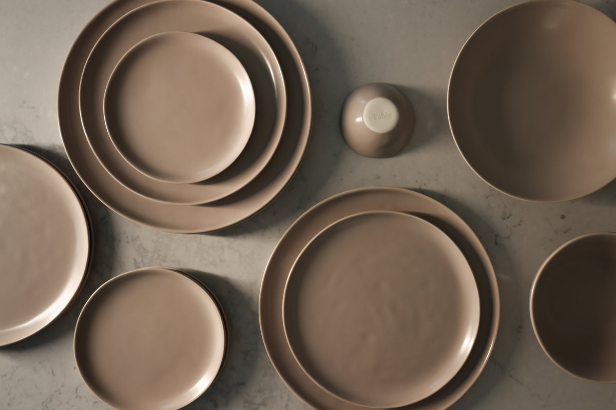Overhead view of Fable matte mauve-taupe stoneware plates and bowls arranged on a marble surface
