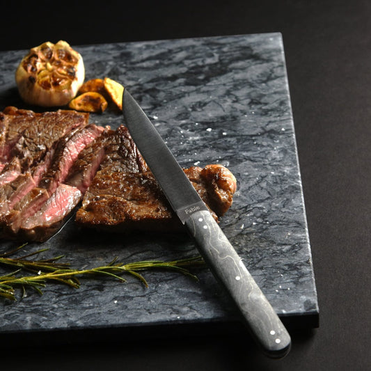 Sliced steak on a dark marble slab with a knife, garlic, and rosemary. #black