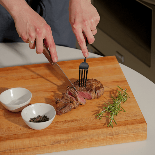 Person cutting a steak on a wooden cutting board with a fork and knife, surrounded by seasonings. #walnut